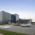 A modern glass office building with the Kellogg’s logo, next to a multi-level parking garage. Two cars drive past on a wide road in the foreground, against a clear sky at dusk.