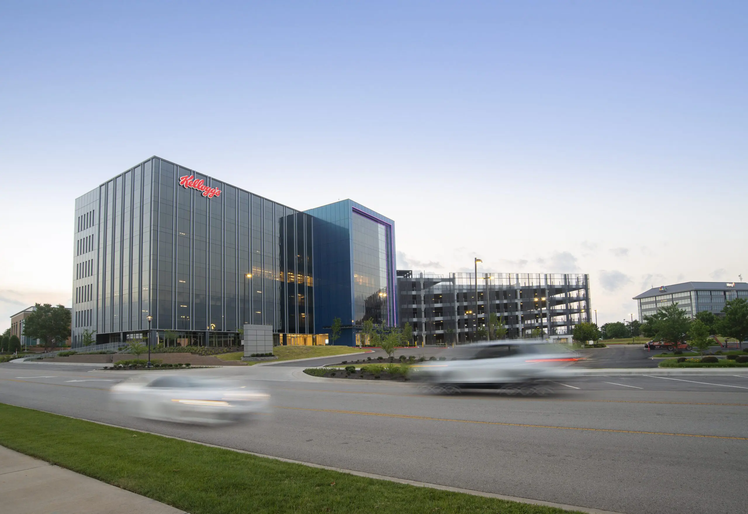A modern glass office building with the Kellogg’s logo, next to a multi-level parking garage. Two cars drive past on a wide road in the foreground, against a clear sky at dusk.
