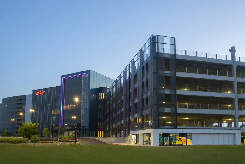 A modern, multi-story office building with glass walls and a Kellogg’s logo, next to a large parking garage, both illuminated at dusk with a clear blue sky in the background.