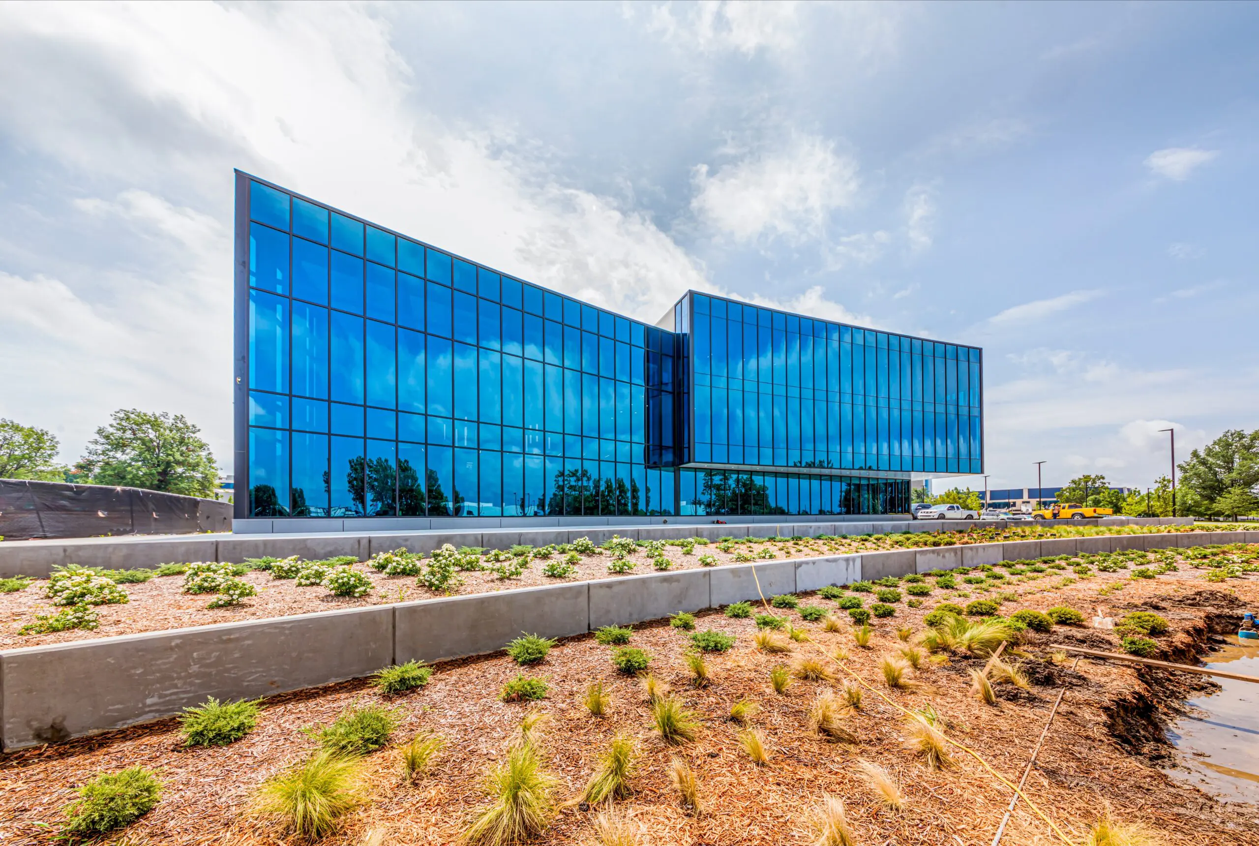 A modern glass office building with reflective blue windows stands behind landscaped grounds with small plants and mulch under a partly cloudy sky.