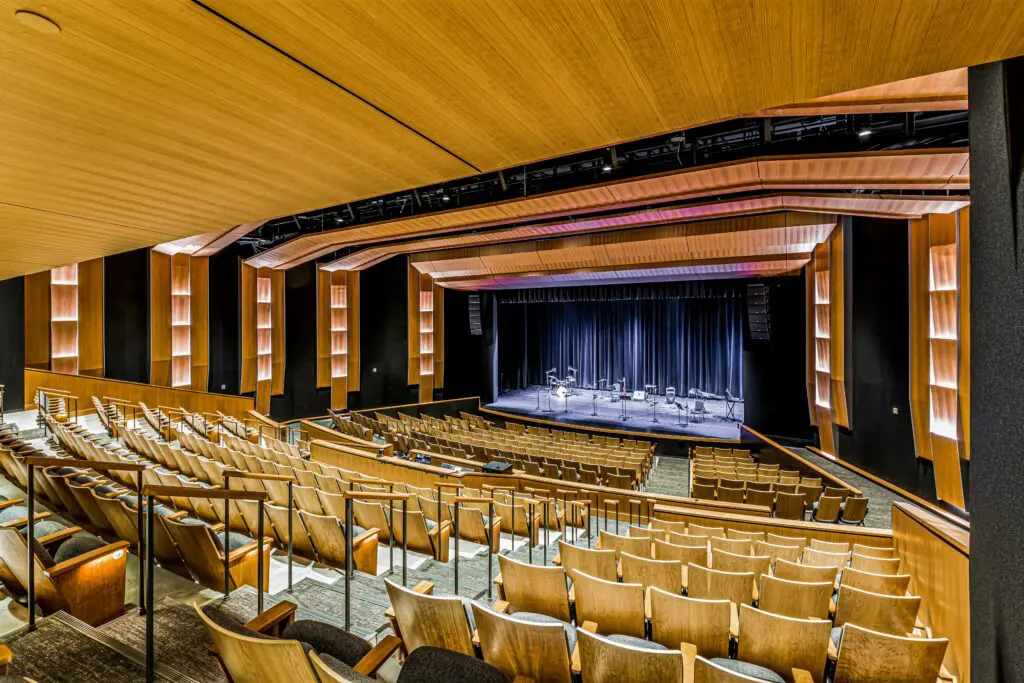 A modern auditorium with rows of wooden seats facing a stage set up with microphones and music stands, surrounded by wooden acoustic panels and soft lighting.
