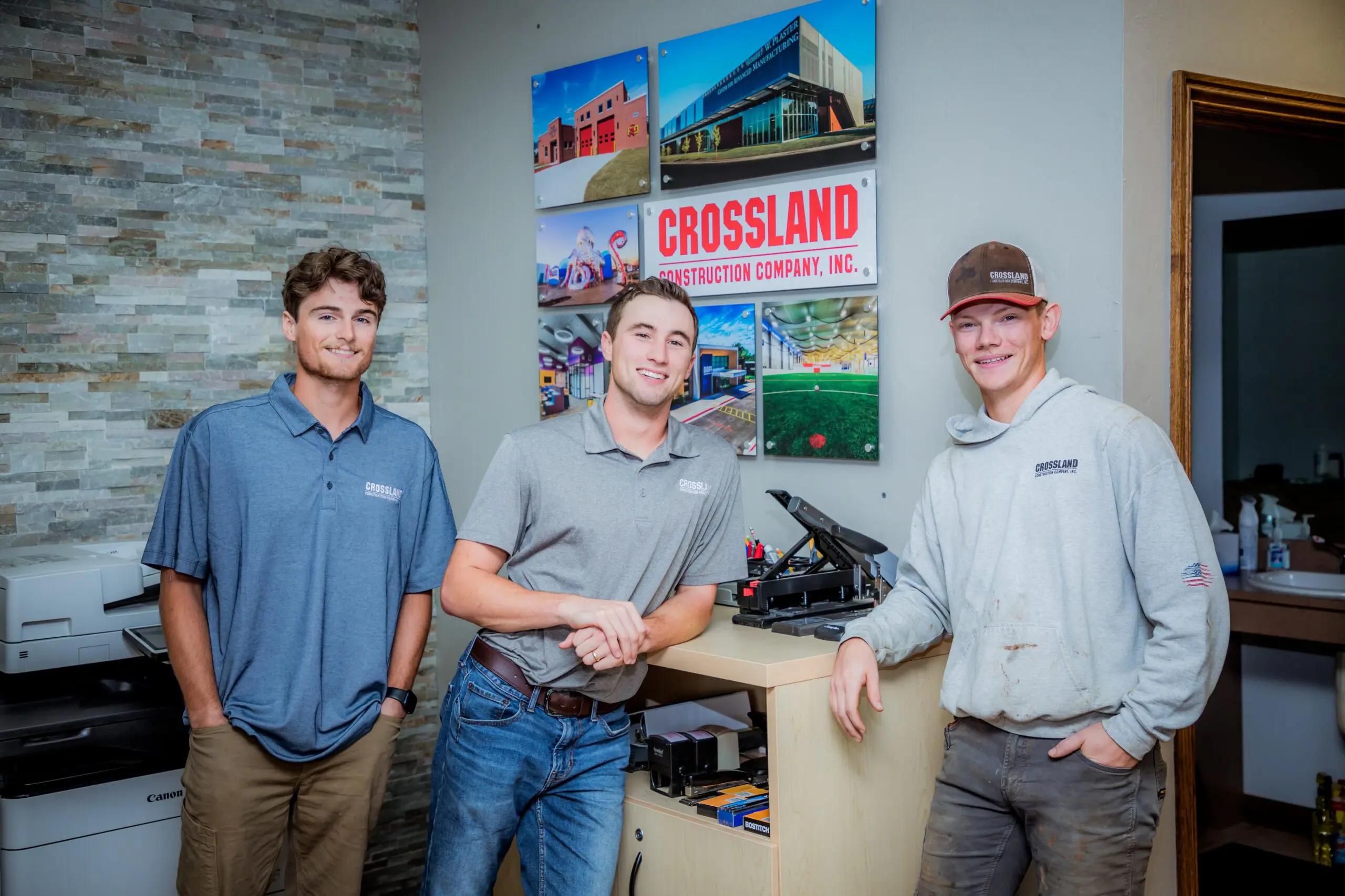 Three young men wearing Crossland Construction shirts stand and smile in an office. Behind them is a wall with Crossland Construction Company signage and project photos. A printer and office supplies are nearby.