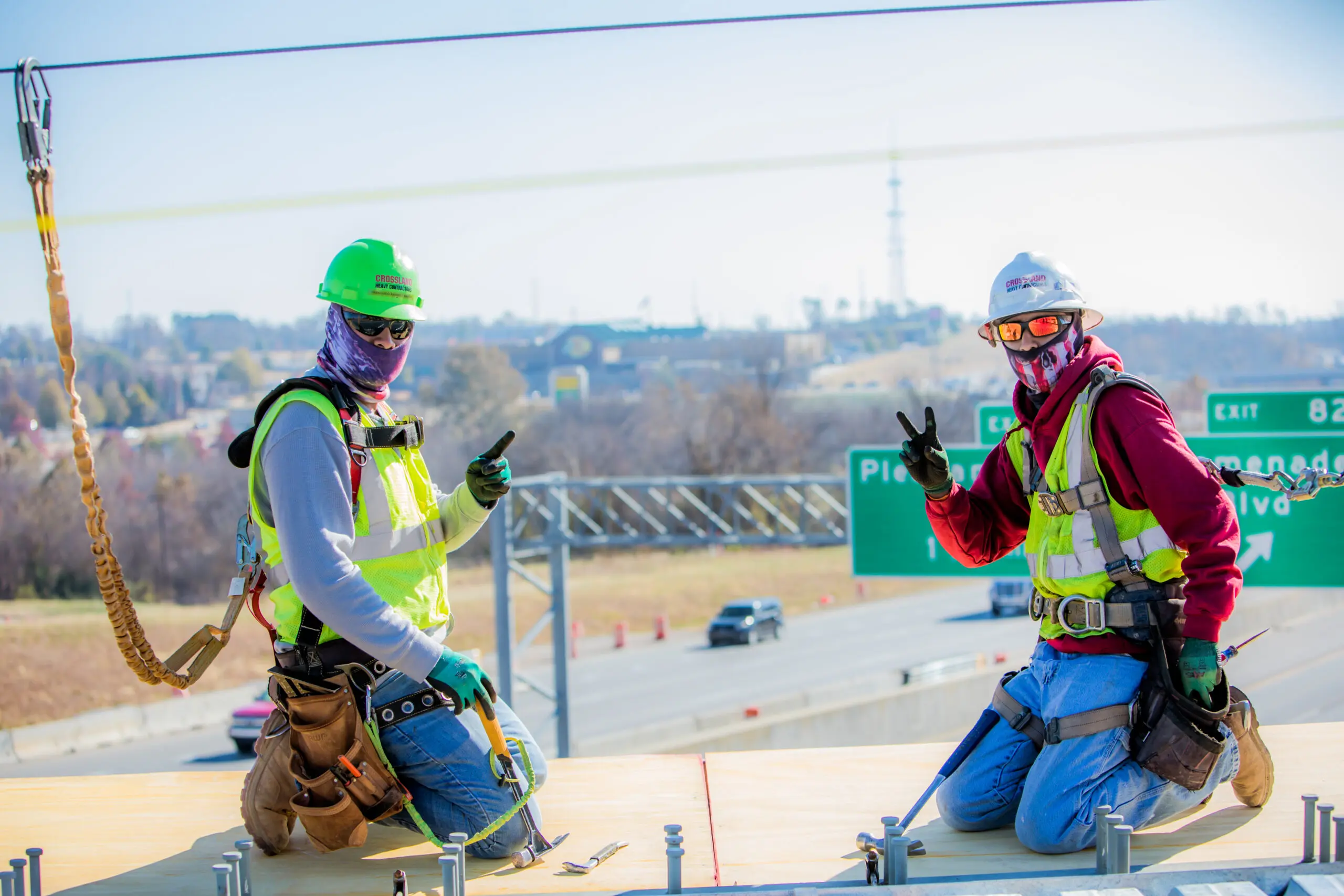 Two construction workers wearing safety gear and masks kneel on a platform, giving peace signs. A highway, cars, and green exit signs are visible in the background under a clear sky.