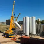 A large yellow crane lifts a tall white cylindrical tank to position it upright beside three similar tanks at an industrial facility on a clear, sunny day.