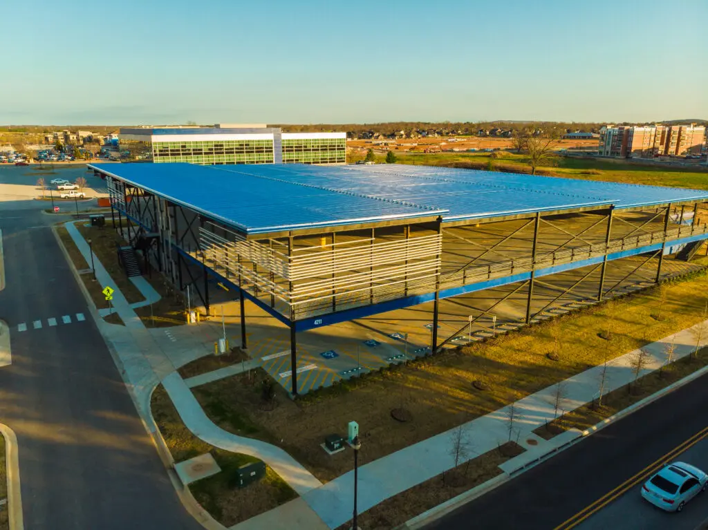 A large building under construction with a blue roof and steel framework, surrounded by roads, sidewalks, and parking areas. Other buildings and open land are visible in the background.