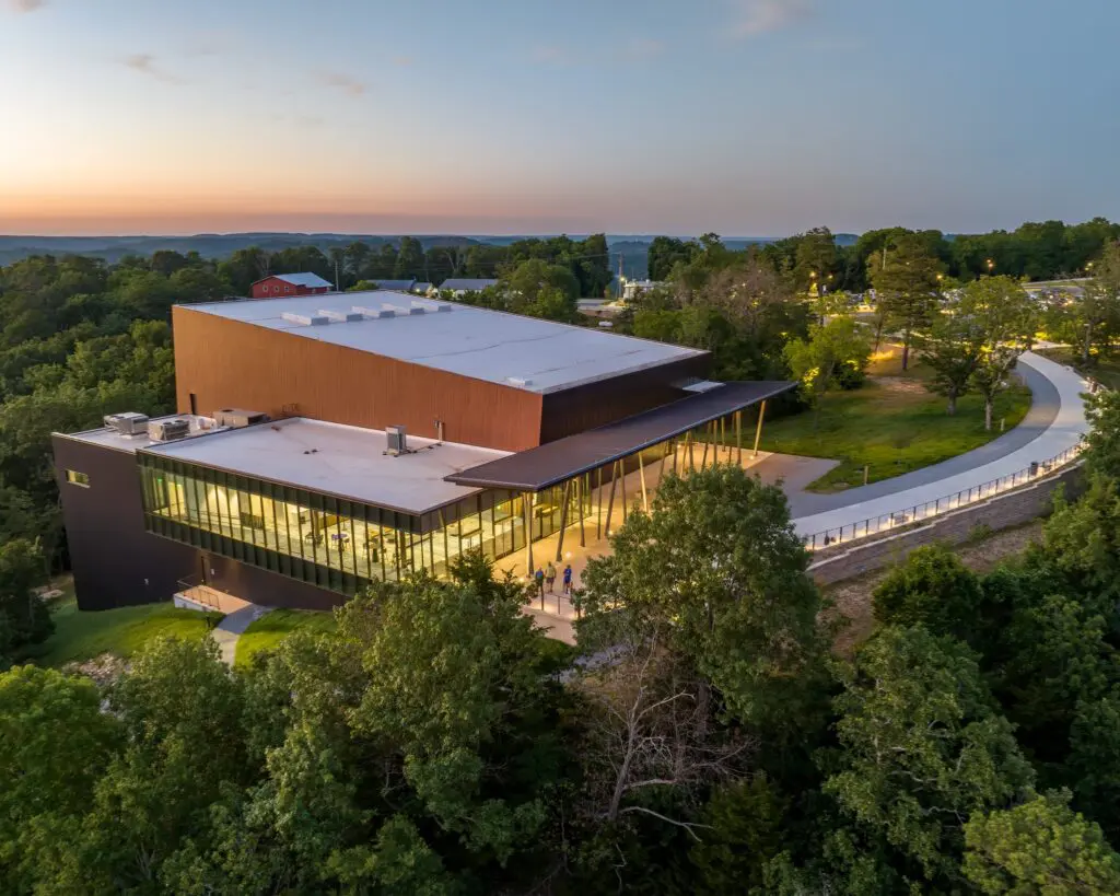 Modern building with large glass windows and a flat roof, surrounded by trees and greenery at sunset. A curved walkway leads to the entrance, and distant hills are visible in the background under a clear sky.