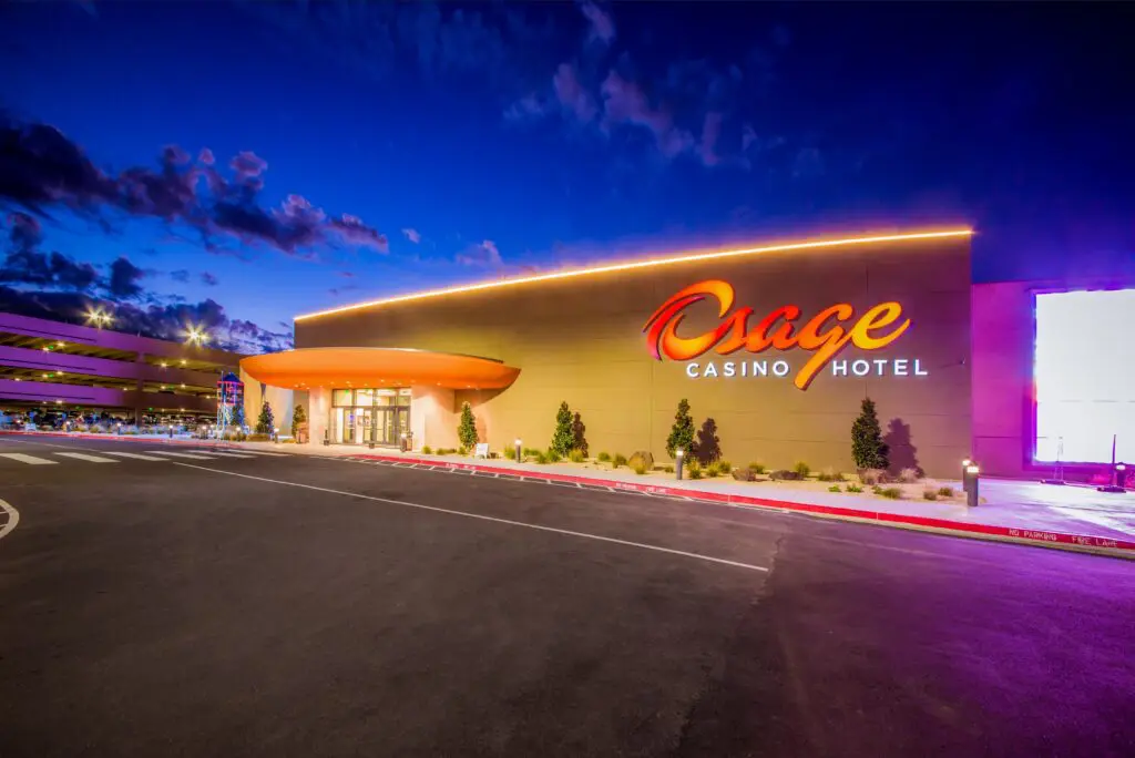 The Osage Casino Hotel building at dusk, illuminated by exterior lights, with its bright orange sign visible and a clear sky in the background. The parking area is empty in front of the entrance.