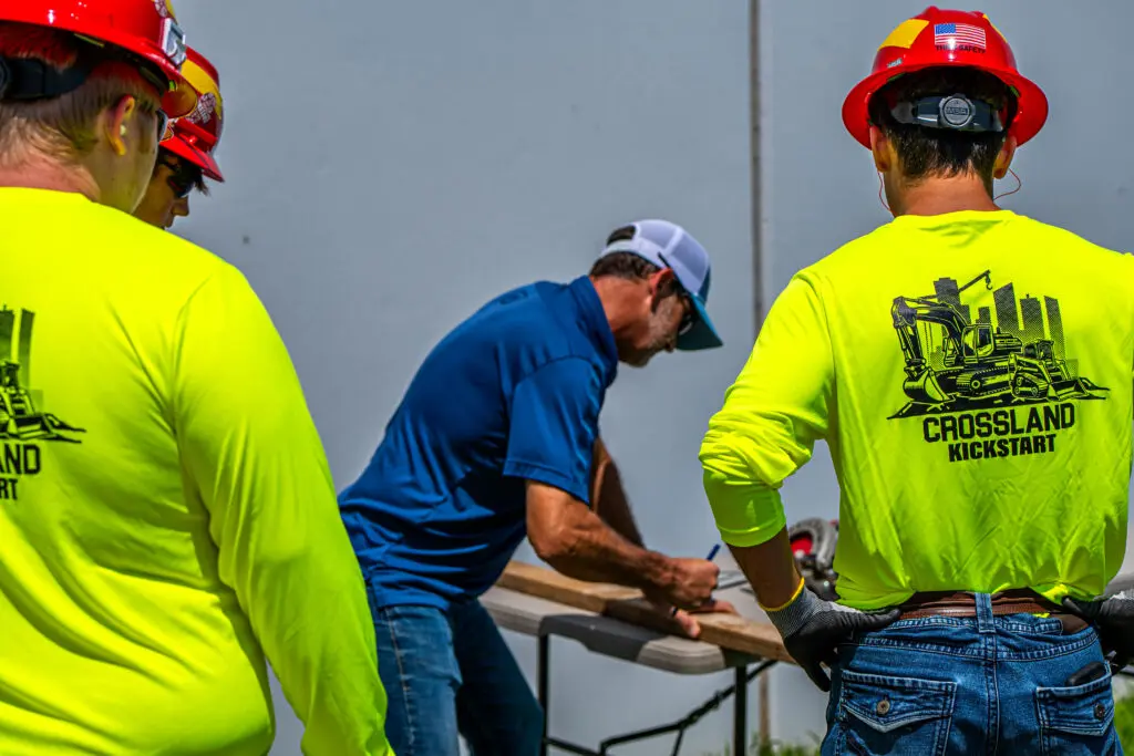 Three construction workers in neon yellow shirts and hard hats watch a man in a blue shirt sign papers on a table outdoors. The shirts have "Crossland Kickstart" and a construction graphic on the back.