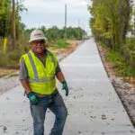 A construction worker in a yellow safety vest and hard hat smiles while standing on a newly paved sidewalk, surrounded by trees and utility poles.