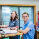 Two people smiling at a standing desk in an office. The woman is typing on a keyboard, and the man is sitting. Papers, office supplies, and a Kansas City Chiefs "Champions" banner are visible in the background.