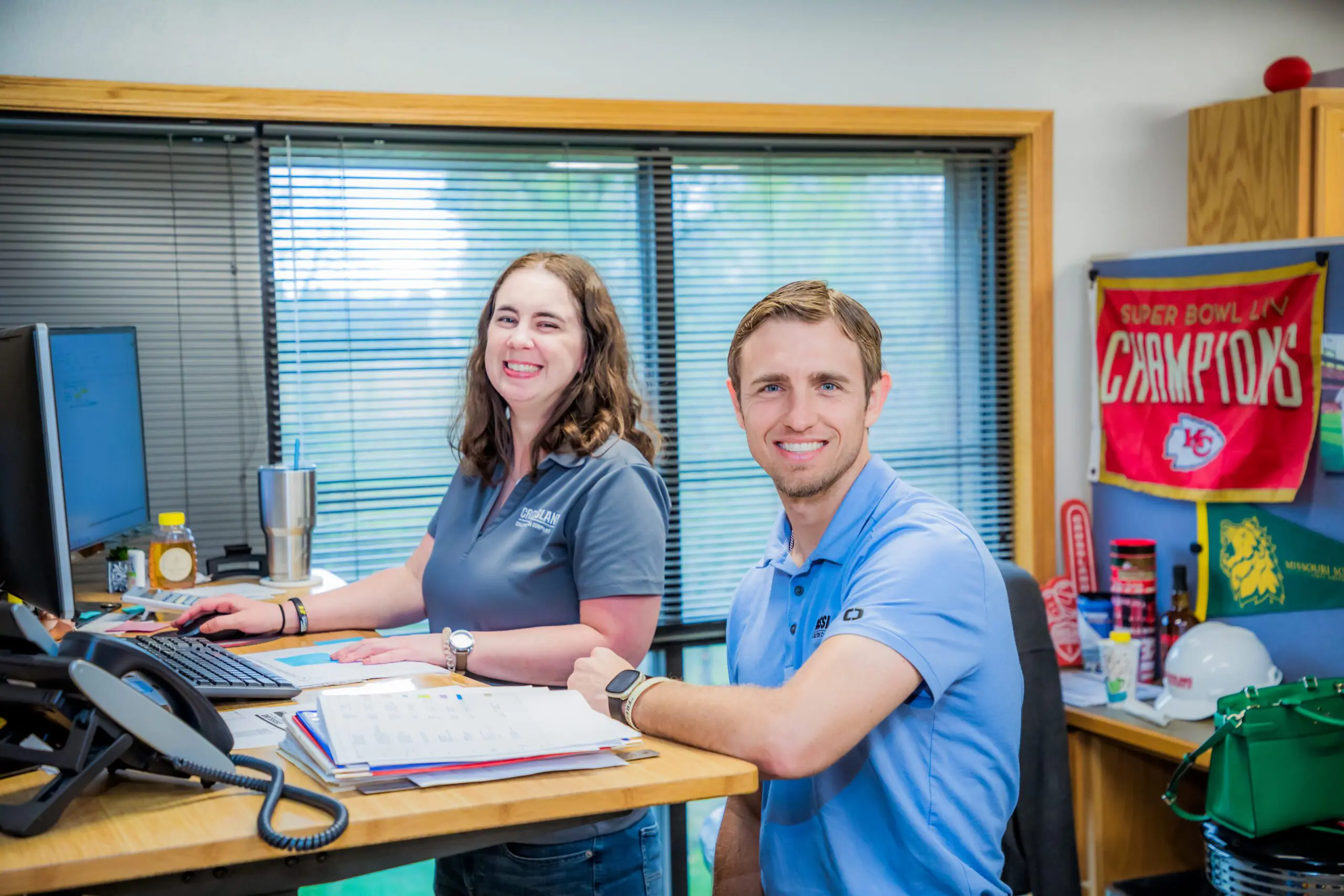 Two people smiling at a standing desk in an office. The woman is typing on a keyboard, and the man is sitting. Papers, office supplies, and a Kansas City Chiefs "Champions" banner are visible in the background.
