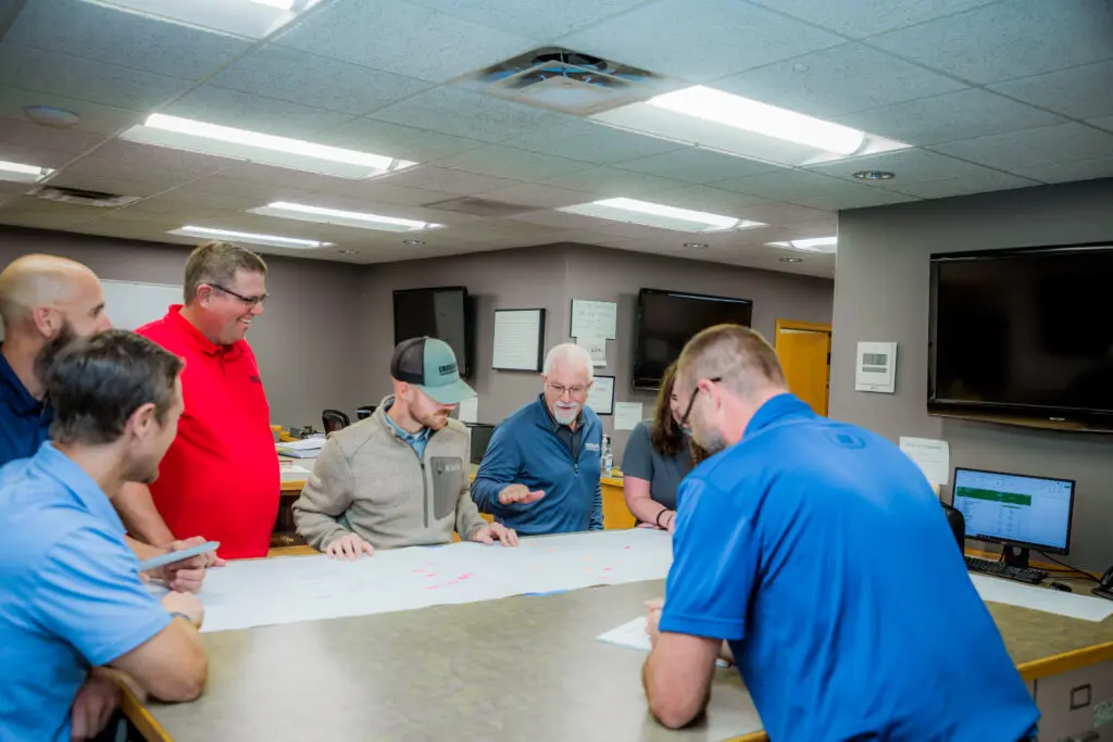 A group of six men gather around a table, looking at large papers spread out, appearing to discuss plans or blueprints in a well-lit office with monitors and bulletin boards on the walls.