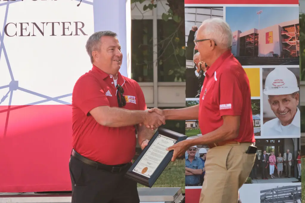 Two men wearing red shirts shake hands; one hands a framed certificate to the other. They stand outside near banners, one showing a building and construction images, the other partially reading "CENTER.