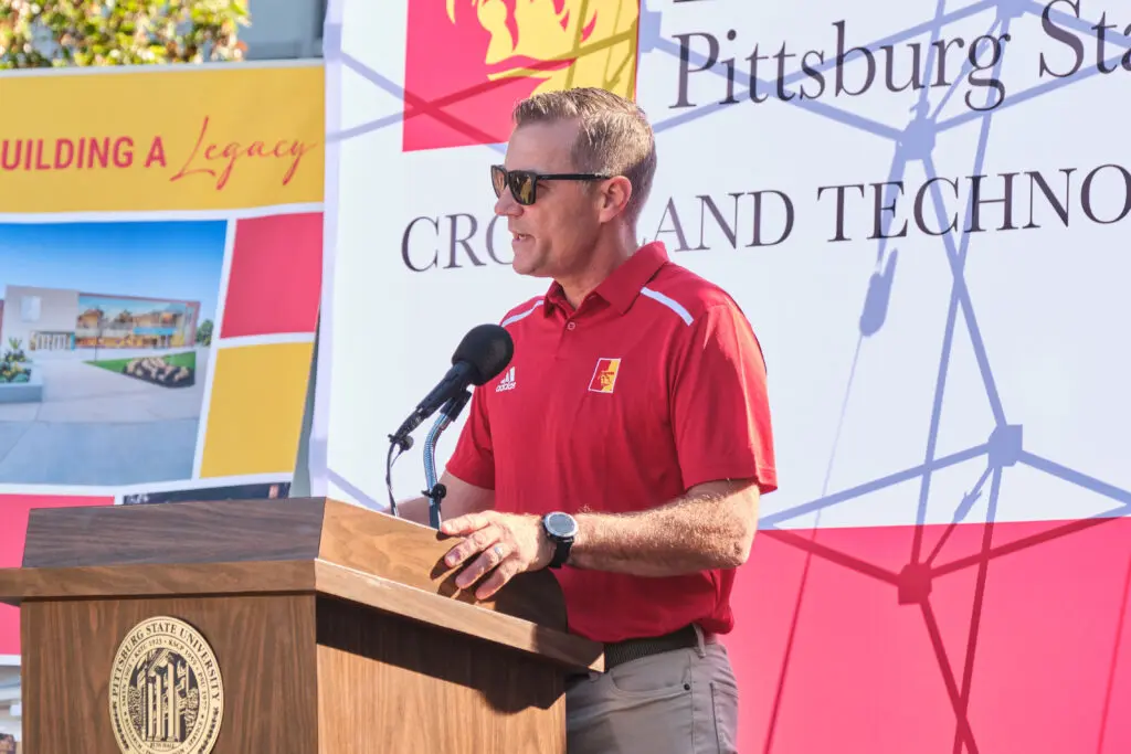 A man in sunglasses and a red Pittsburg State University polo shirt speaks at a podium with a microphone. Behind him are banners for Pittsburg State University and a "Building a Legacy" sign.