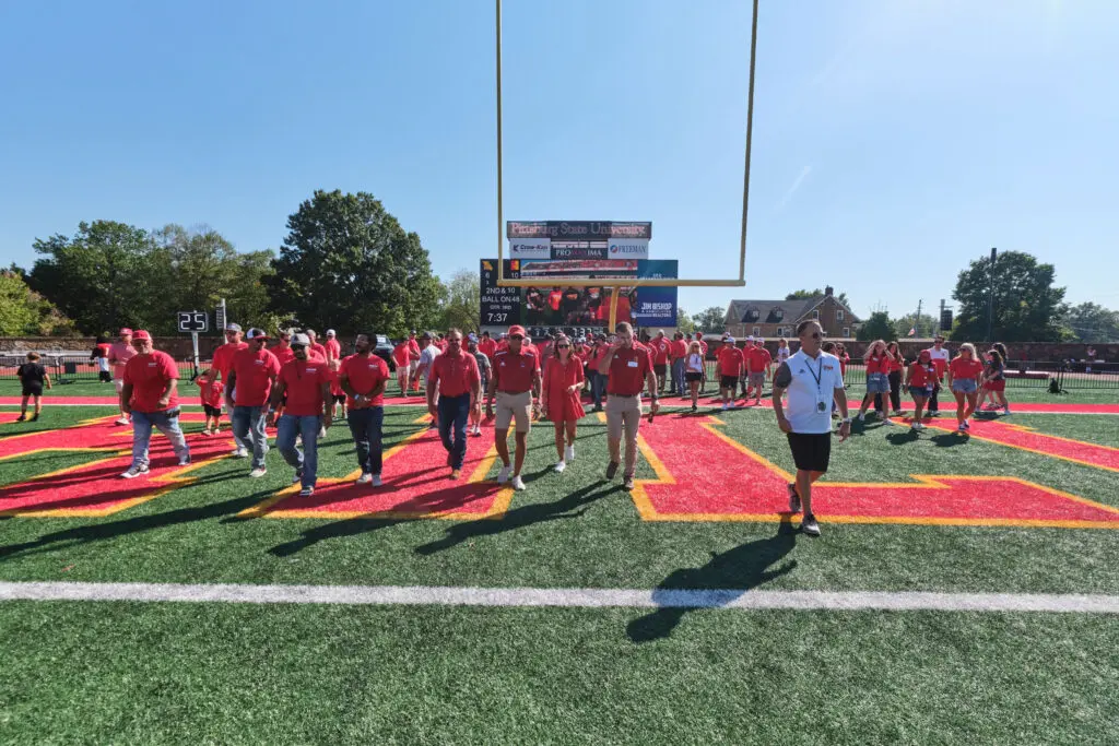 A large group of people wearing red shirts walk across a football field toward the camera, with a scoreboard and goalpost visible in the background on a sunny day. One person in white stands to the right.