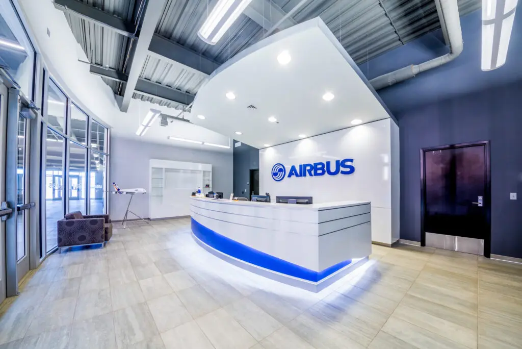 A modern, brightly-lit office lobby with a curved reception desk displaying the Airbus logo. The space has light wood flooring, gray walls, and exposed ceiling pipes. A single armchair sits near the glass entry doors.