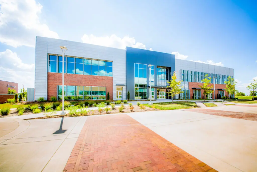 A modern two-story building with large windows, brick and metal exterior, surrounded by landscaped greenery and a wide paved walkway under a bright blue sky with scattered clouds.