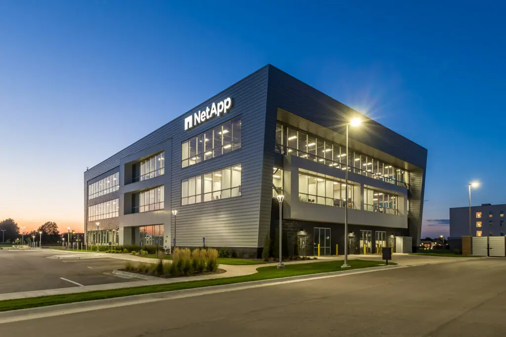 A modern three-story building with large windows and "NetApp" signage, pictured at dusk with interior lights on and empty parking spaces in front. Streetlights illuminate the scene.