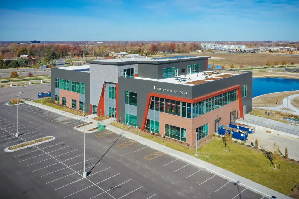 A modern two-story building labeled "The Smart Factory," featuring large windows and red brick and gray paneling, is surrounded by an empty parking lot and landscaped areas under a clear sky.