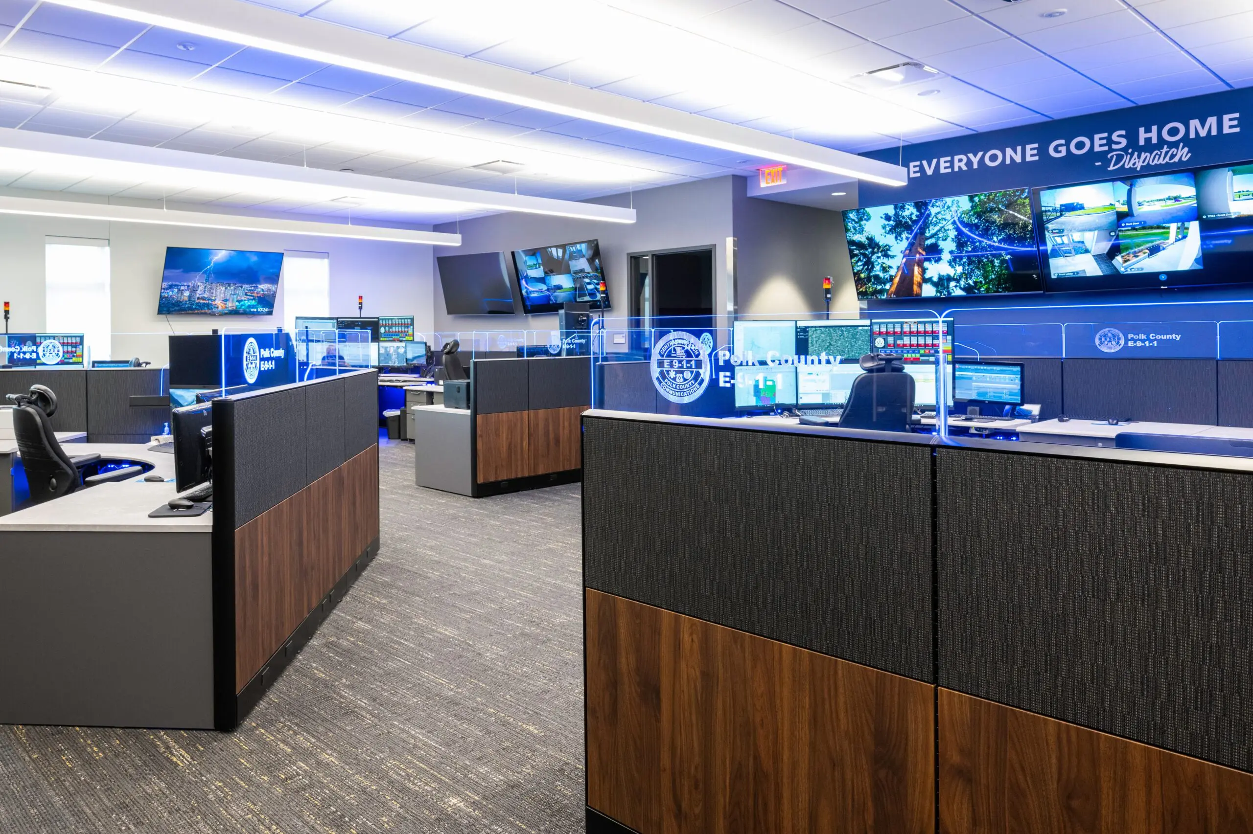 A modern emergency dispatch center with multiple workstations, large monitors displaying maps and security footage, and a sign reading “EVERYONE GOES HOME – Dispatch” on the wall. The room is brightly lit and organized.