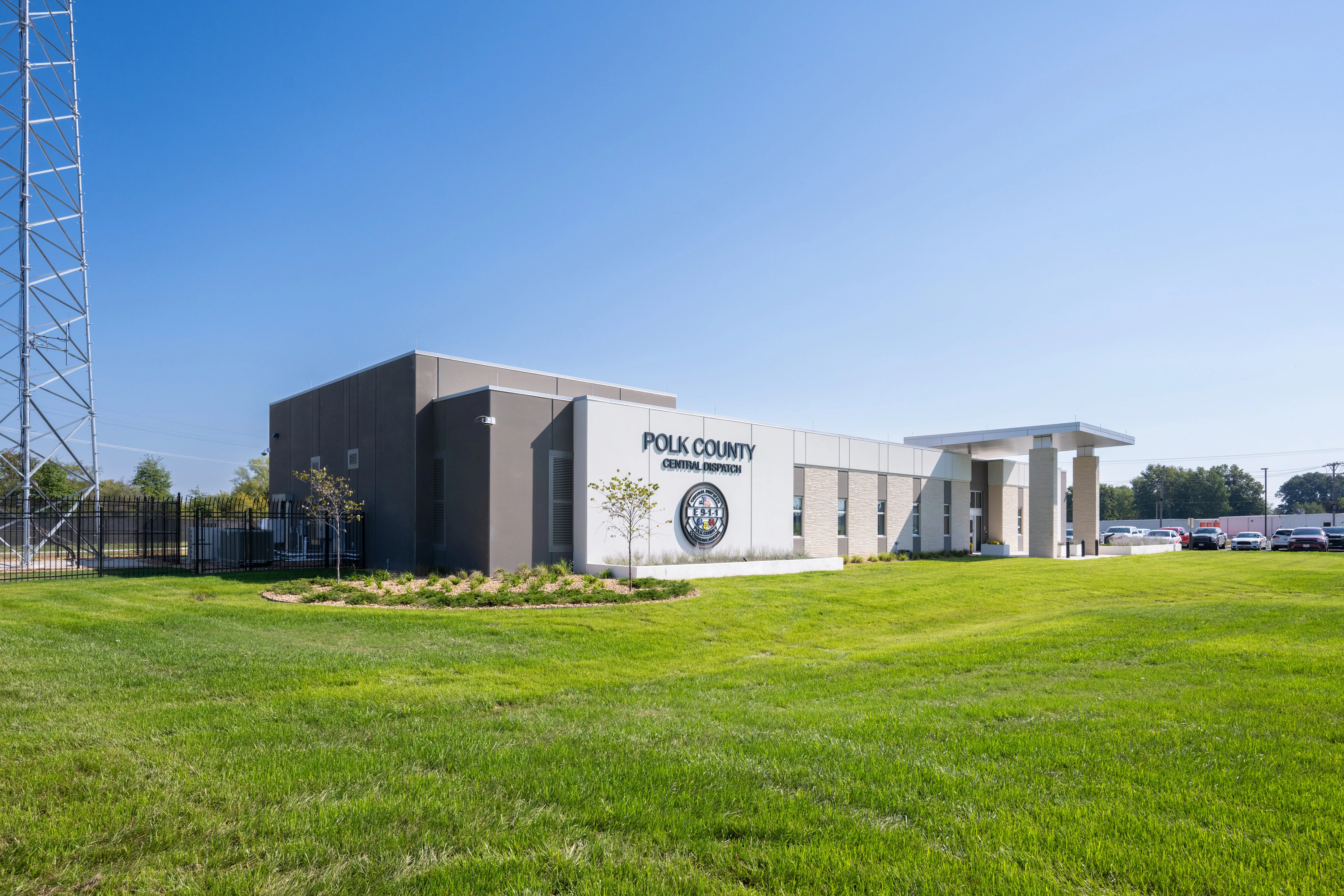 A modern, single-story building labeled "Polk County Central Dispatch" sits on a well-maintained grassy lawn under a clear blue sky. A few small trees and parked cars are visible nearby.