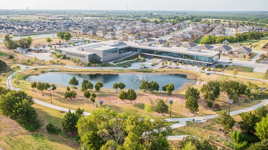 Aerial view of a modern building with a pond and fountain in front, surrounded by trees, walking trails, and a suburban neighborhood with houses in the background under a clear sky.