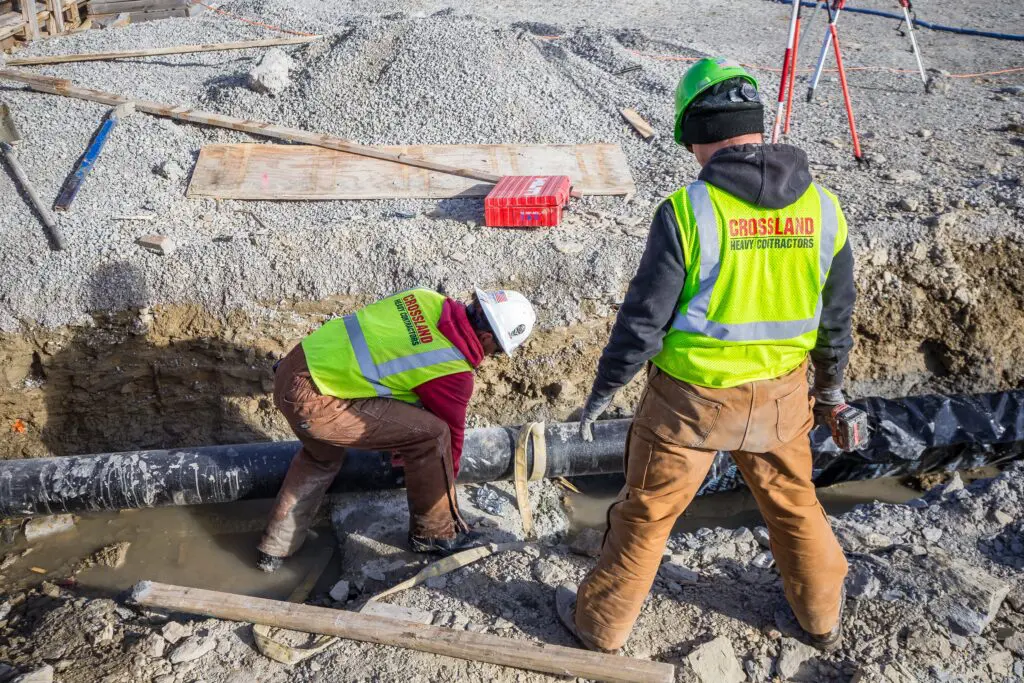 Two construction workers in safety vests and helmets work on a large black pipe in a muddy trench at a construction site, surrounded by gravel and wooden planks.