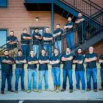 Sixteen men in matching black shirts and jeans stand with arms crossed in front of a building with stairs. Some stand on the ground, others on the stairs, posing for a group photo outdoors.