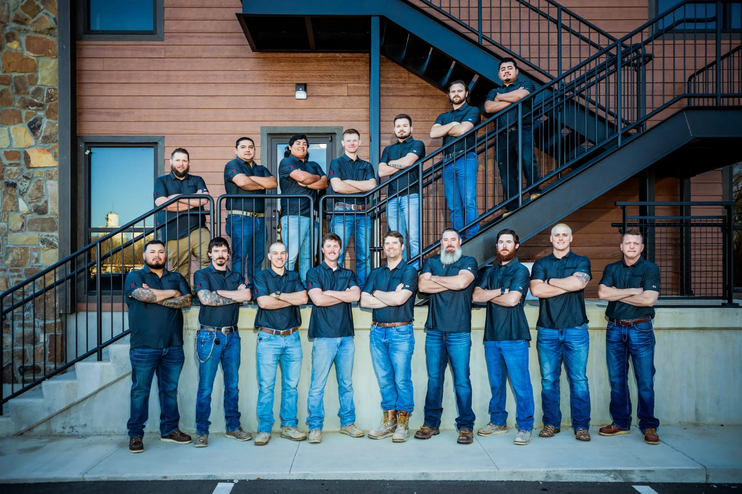 Sixteen men in matching black shirts and jeans stand with arms crossed in front of a building with stairs. Some stand on the ground, others on the stairs, posing for a group photo outdoors.