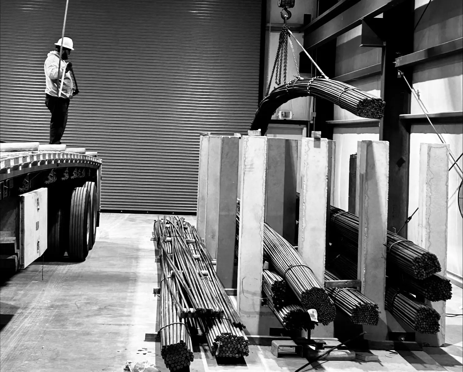 A worker in a hard hat stands on a flatbed trailer, overseeing steel rods being lifted by a crane inside an industrial warehouse. Concrete barriers and bundles of rods are stacked nearby. Black and white image.