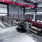 Metal fabrication workshop with industrial machinery, including a red RMS saw, metal bars, and partially assembled structures. The area is spacious with concrete floors and exposed metal beams.