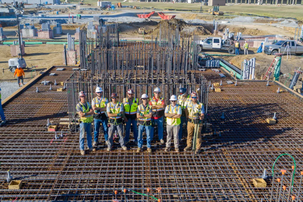 Seven construction workers stand on a large, rebar-covered building foundation at a construction site, wearing safety vests and hard hats, with construction equipment and vehicles in the background.