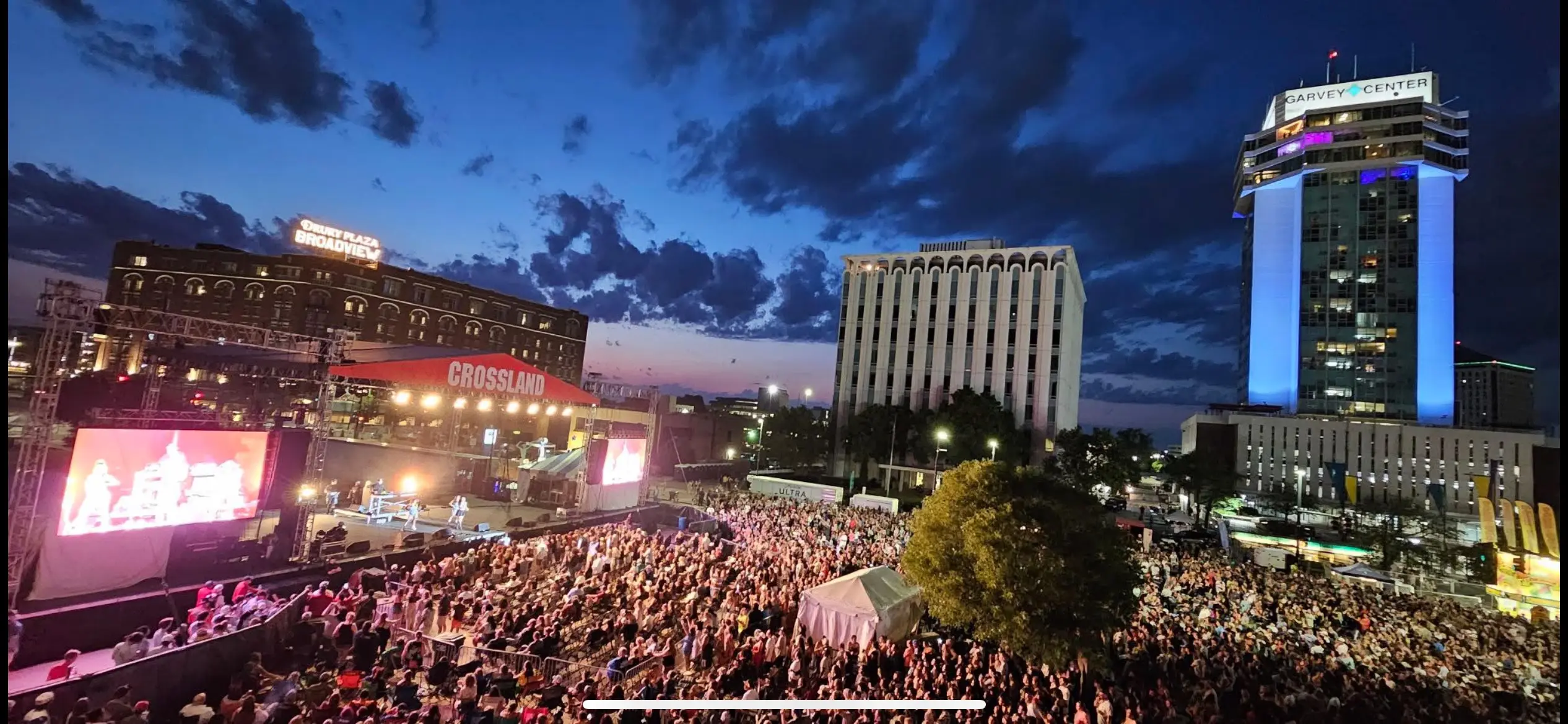 A large crowd gathers at an outdoor concert in a city plaza at dusk, with stage lights illuminating performers and screen displays, surrounded by tall, lit-up buildings under a dramatic evening sky.