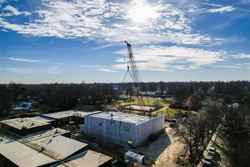 A large crane stands beside a partially constructed building under a bright, partly cloudy sky. Surrounding the site are trees, houses, and open land, with sunlight shining overhead.