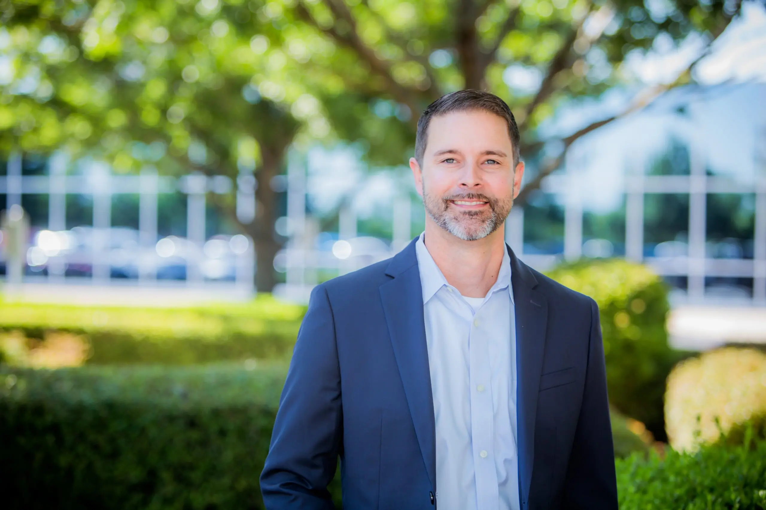 A man in a navy suit and light blue shirt stands outdoors in front of green bushes and trees, smiling at the camera. The background is bright and features blurred greenery and building windows.