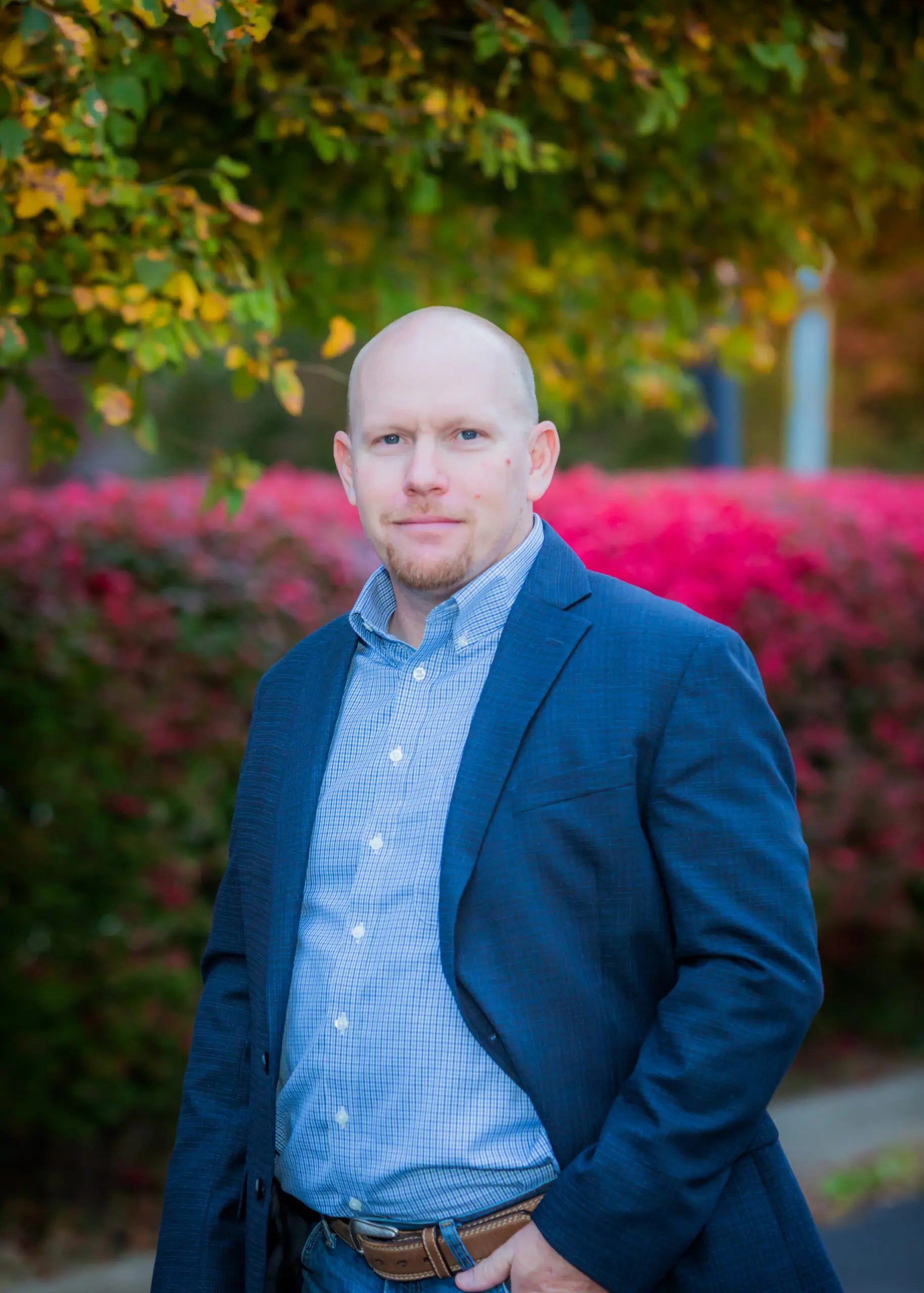 A man in a blue blazer and checked shirt stands outdoors in front of bright pink and green foliage, posing with one hand in his pocket and a neutral expression.