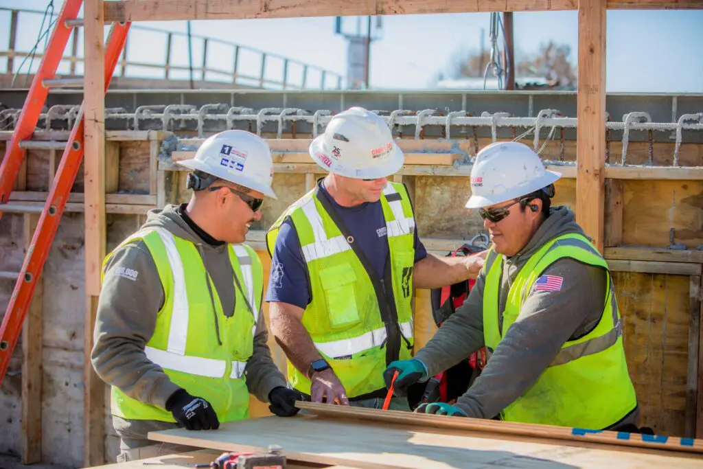 Three construction workers wearing safety helmets and neon vests discuss blueprints at a wooden table on a building site, with tools and a ladder visible in the background.
