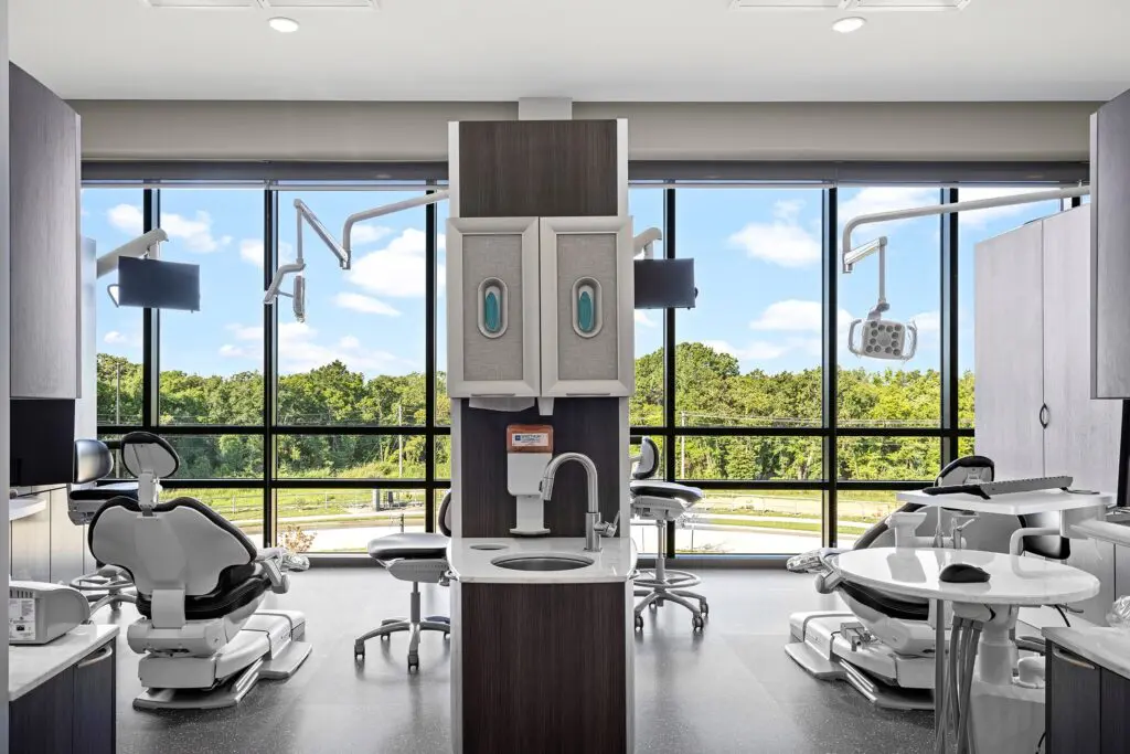 Modern dental office with multiple dental chairs, equipment, and sinks, set by large windows overlooking a green landscape under a blue sky with clouds. The space is clean, bright, and organized.