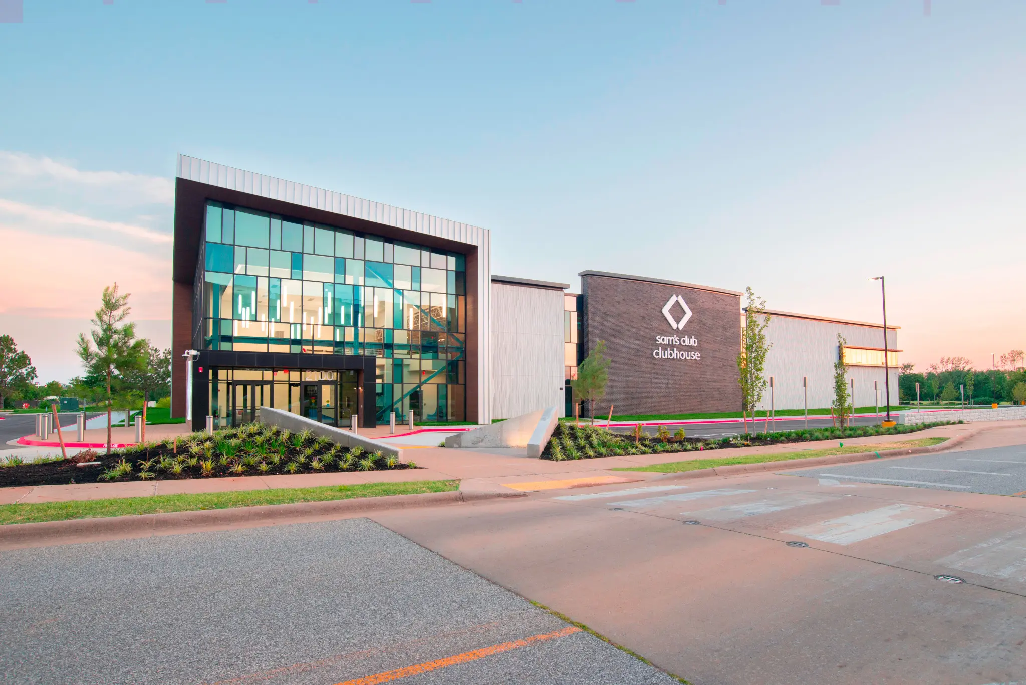 Modern building with large glass windows and the words "Sam's Club clubhouse" on the exterior, surrounded by landscaped greenery and a wide sidewalk at sunset.