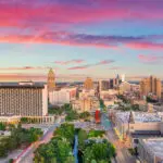 Aerial view of downtown San Antonio, Texas at sunrise, featuring tall buildings, green trees, and a colorful sky with pink and blue clouds.