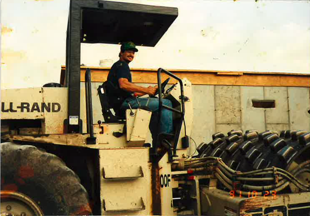 A man wearing a cap and jeans sits and smiles on the seat of a large construction vehicle in an industrial area, with tires and a concrete wall visible in the background.