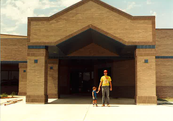 An adult and a child holding hands stand in front of a large, tan brick building with a geometric entrance under a sunny sky.