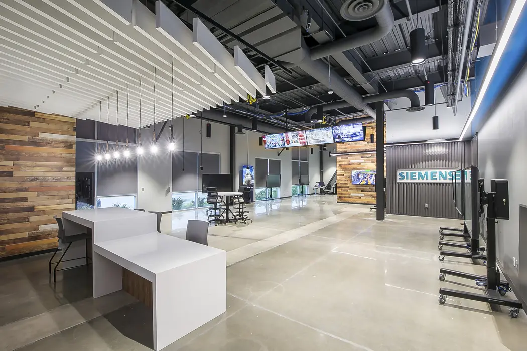 Modern office space with sleek white desks, polished concrete floors, wood accent walls, hanging lights, multiple monitors, and a Siemens sign on a large wall. Ceiling features exposed ductwork and beams.