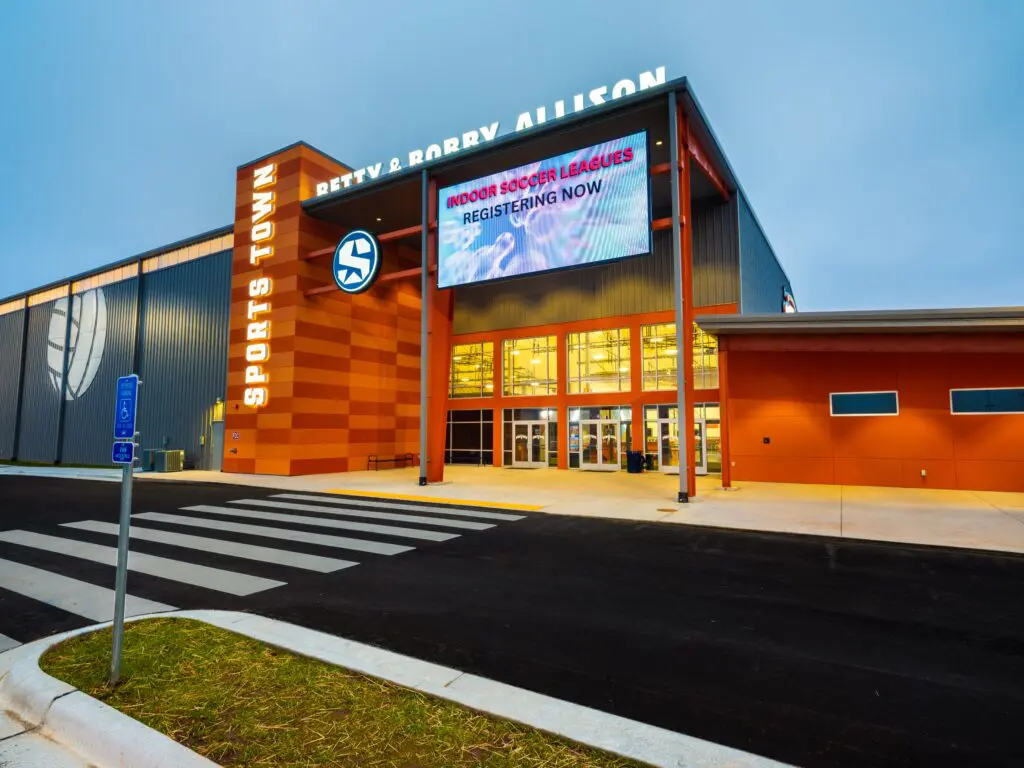 A modern sports facility with a glass entrance, large digital sign advertising indoor soccer leagues registration, and "Sports Town" and "Betty & Bobby Allison" displayed on the building.