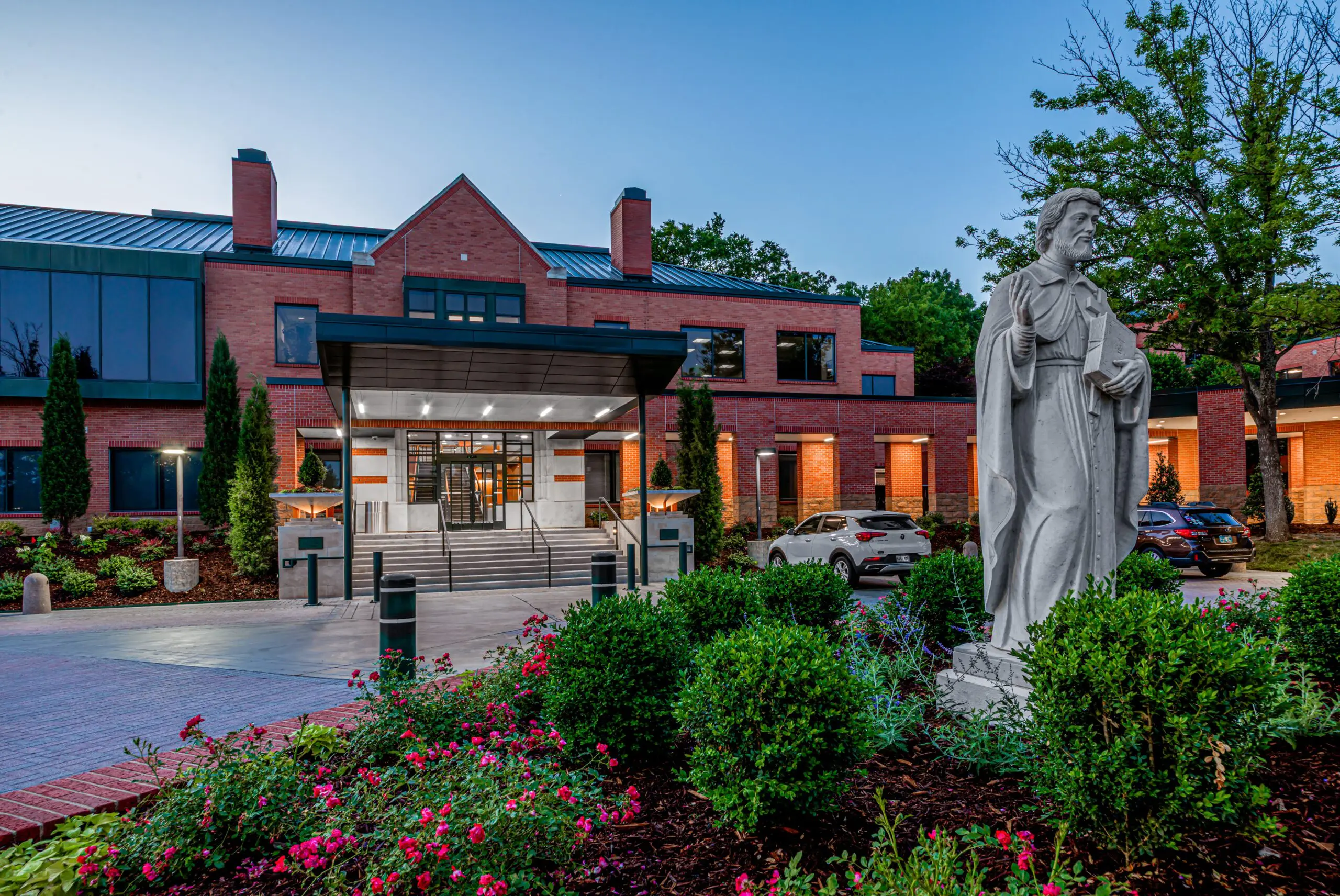 A red-brick building with large windows and a covered entrance is surrounded by landscaped greenery and pink flowers. A stone statue of a robed figure stands in the foreground near parked cars and trees at dusk.