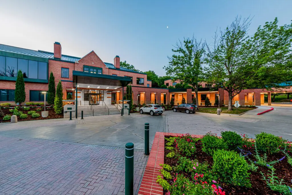 A modern brick building with large windows and a covered entrance, surrounded by neatly landscaped gardens, trees, and parked cars at dusk.