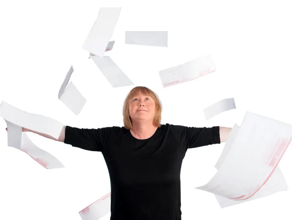 A person with shoulder-length blonde hair wearing a black shirt stands with arms outstretched, looking up as papers fly in the air around them against a white background.