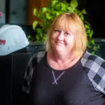 A woman with blonde hair and a gray plaid-sleeved top smiles while standing indoors. A white hard hat with red text and a leafy green plant are visible in the background.