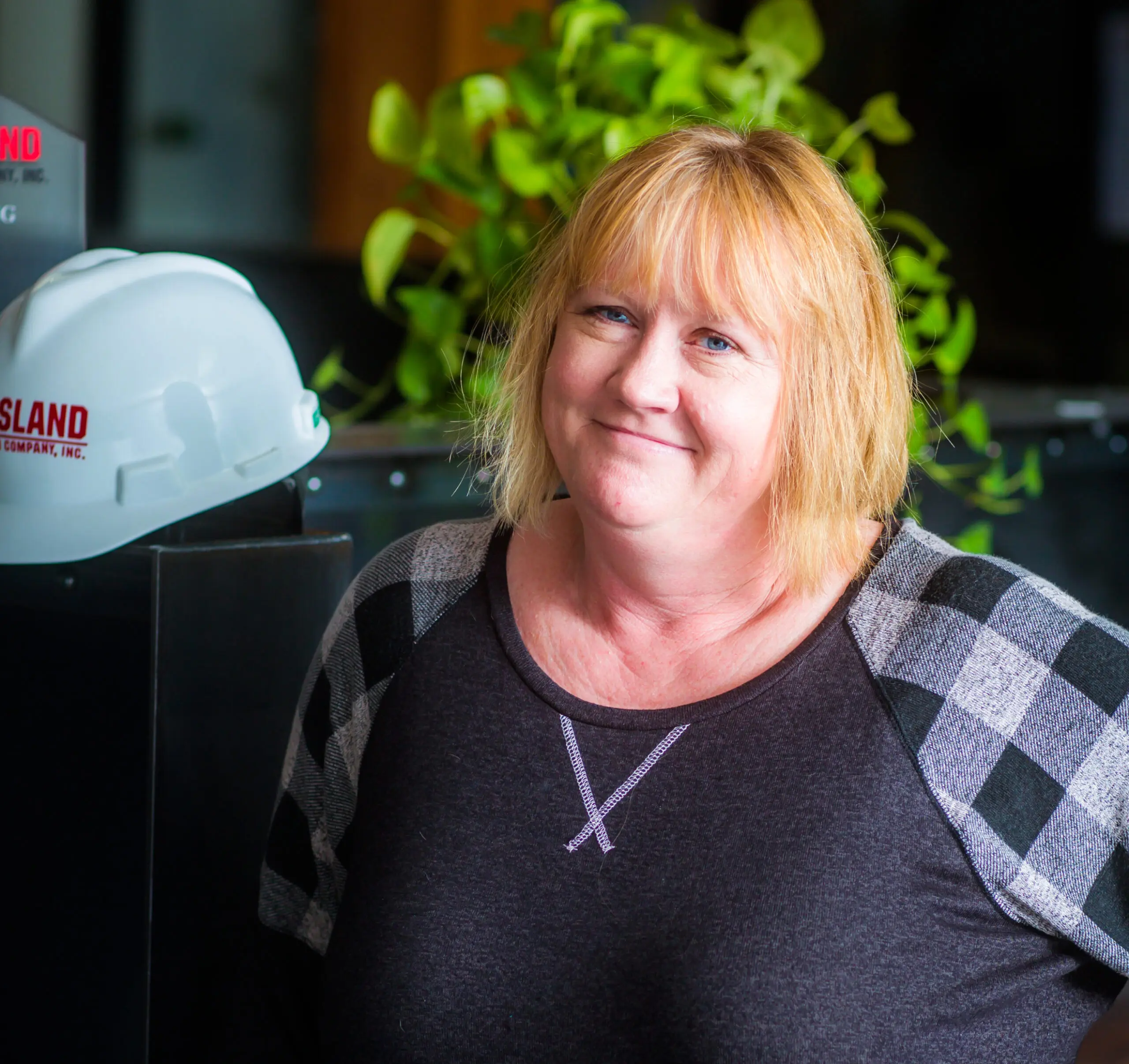 A woman with blonde hair and a gray plaid-sleeved top smiles while standing indoors. A white hard hat with red text and a leafy green plant are visible in the background.