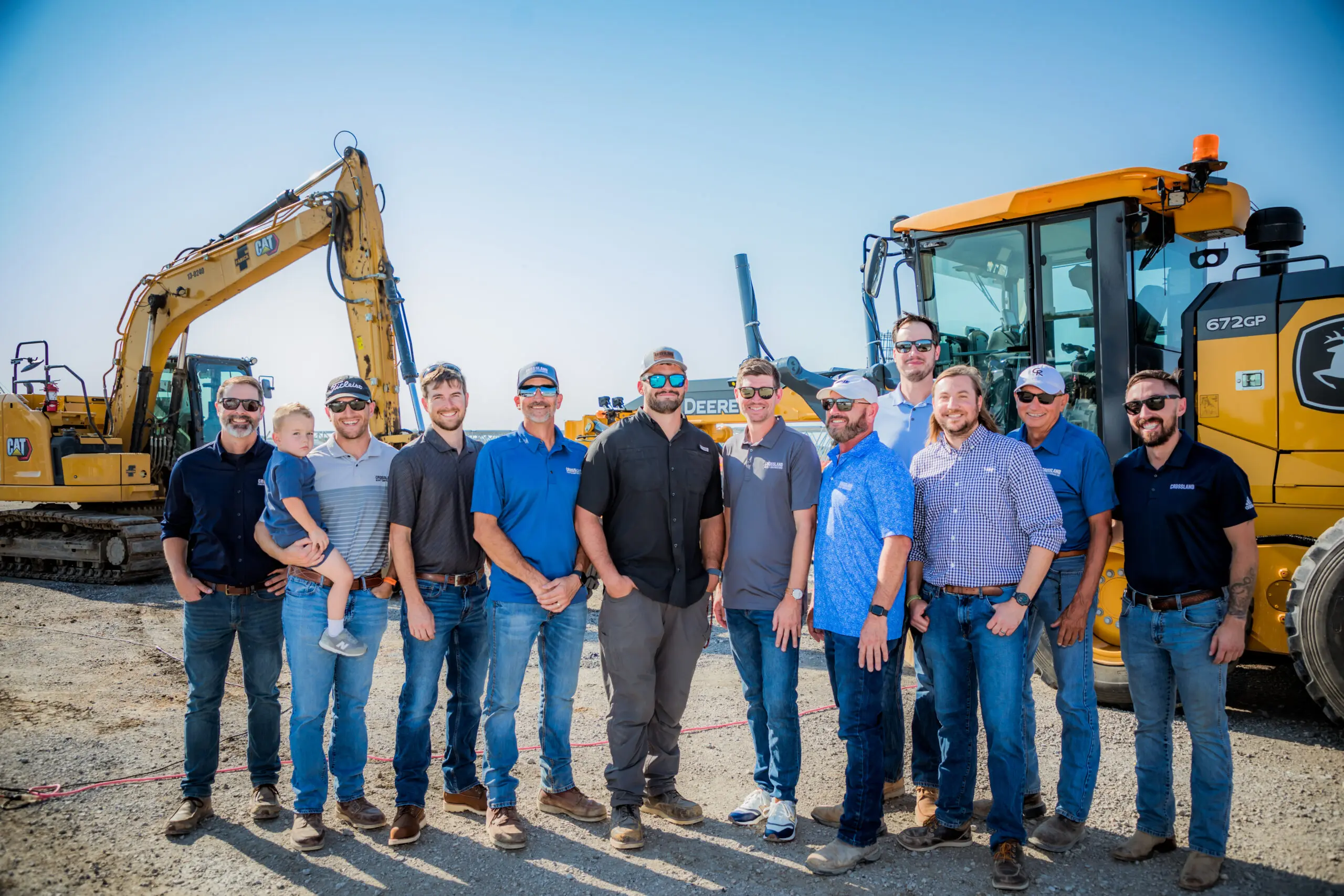 A group of eleven men and a young child stand together outdoors in front of large construction vehicles, wearing casual shirts, jeans, hats, and sunglasses, smiling on a sunny day at a construction site.